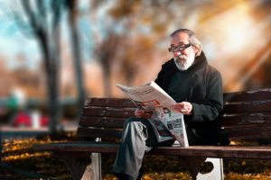 Senior man with beard reading a newspaper on a wooden bench in a sunny park setting.